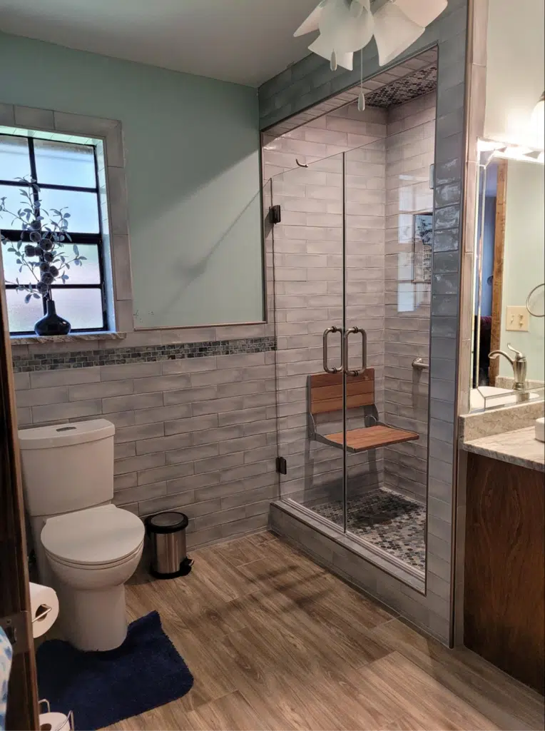 Contemporary bathroom with glass-enclosed shower, wooden bench, and light gray wall tiles.